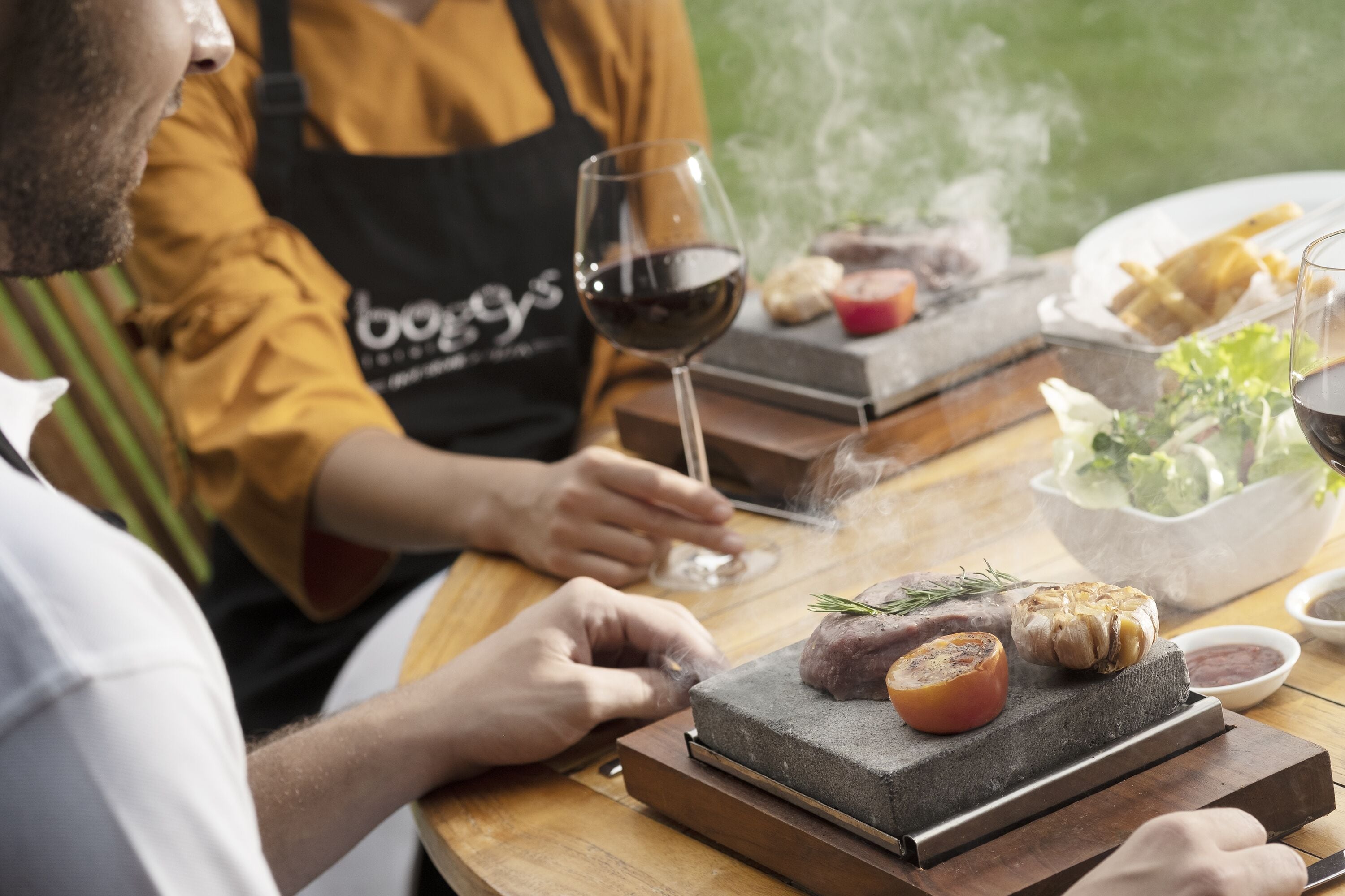 A person grilling meat on a stone slab with a glass of red wine and a bowl of salad on the table.