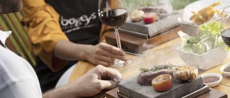 A person grilling meat on a stone slab with a glass of red wine and a bowl of salad on the table.