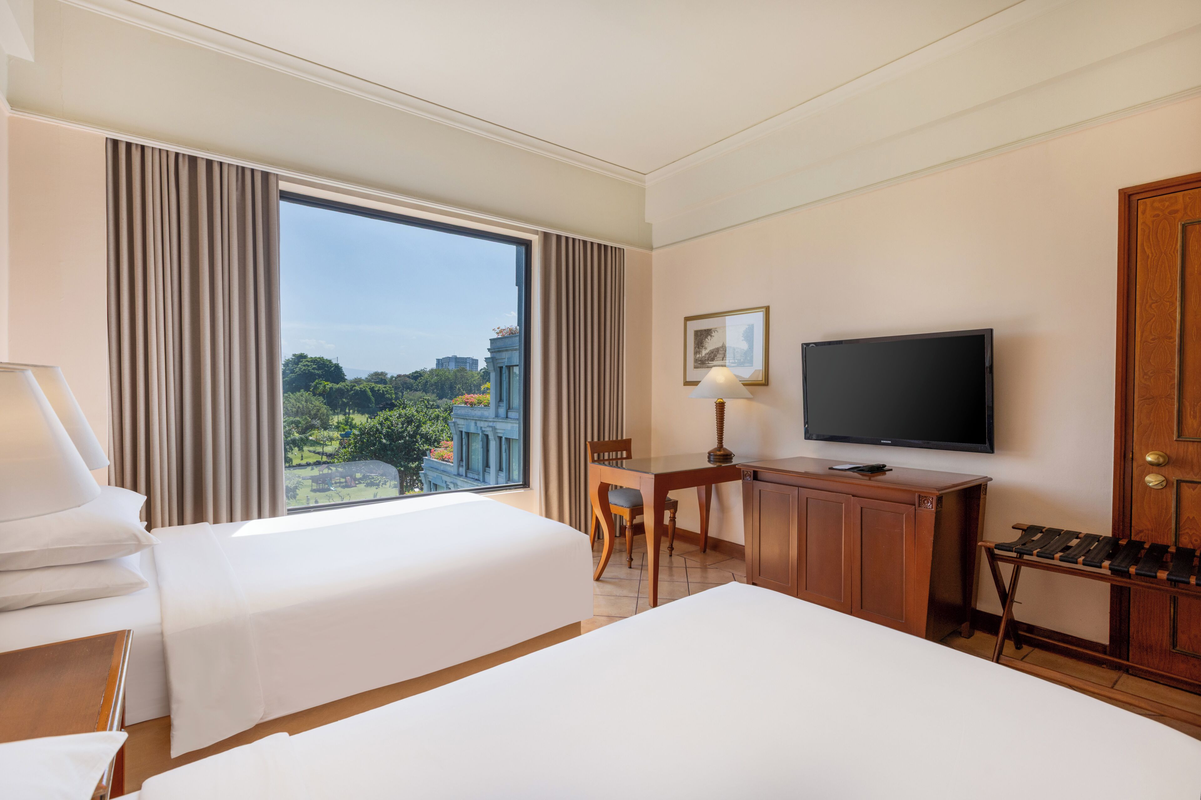 Habitación de hotel con dos camas, un escritorio, una silla, un televisor y un ventanal con vistas a la ciudad.