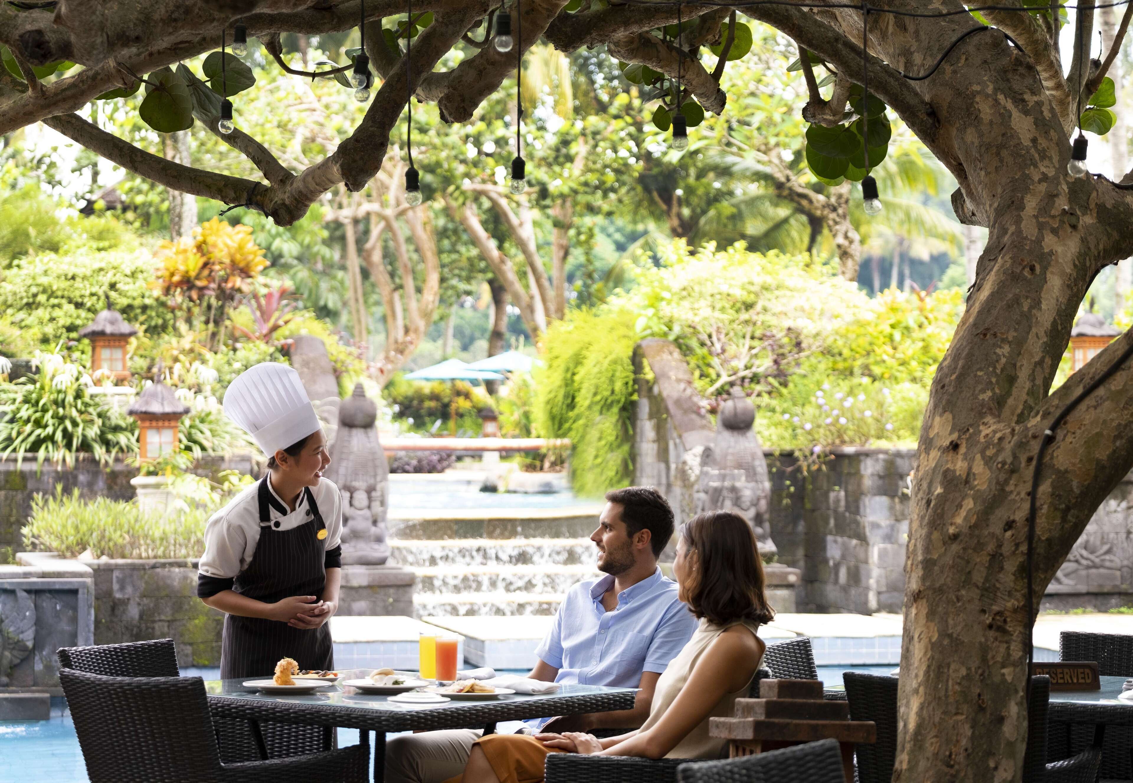 A dining area with wicker furniture, a table with plates and a drink, and a waiter in uniform.