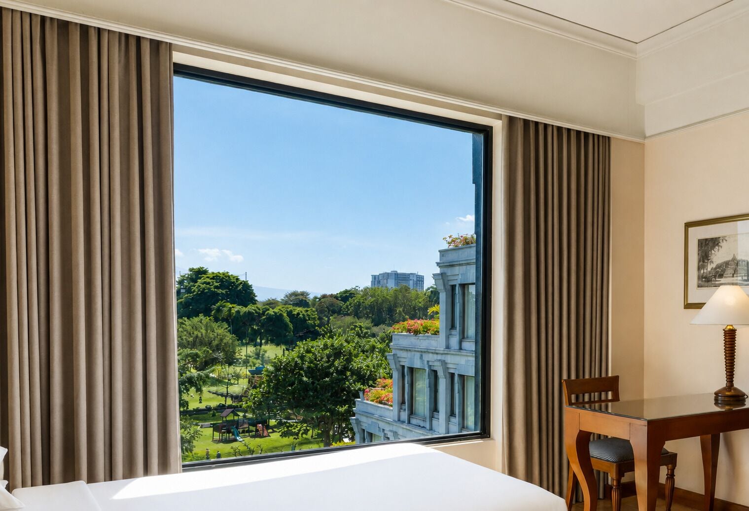A hotel room with a large window, a bed, a desk with a chair, and a lamp.