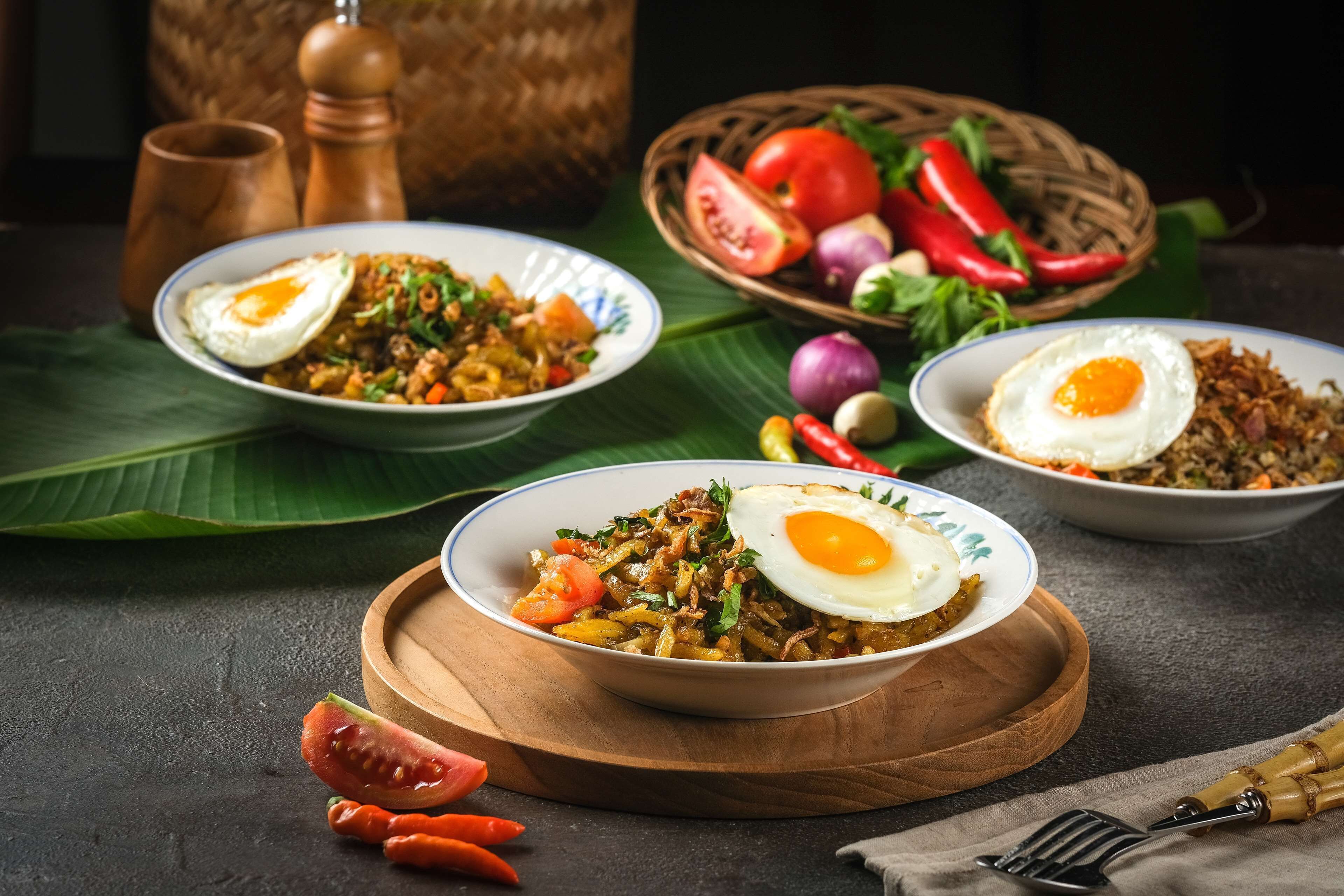 A table with two bowls of fried rice topped with fried eggs, surrounded by fresh vegetables and a wooden board.