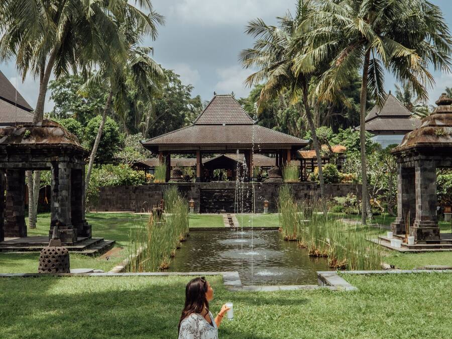 A woman sitting on a checkered blanket with a picnic setup in a lush garden with palm trees and a water feature.