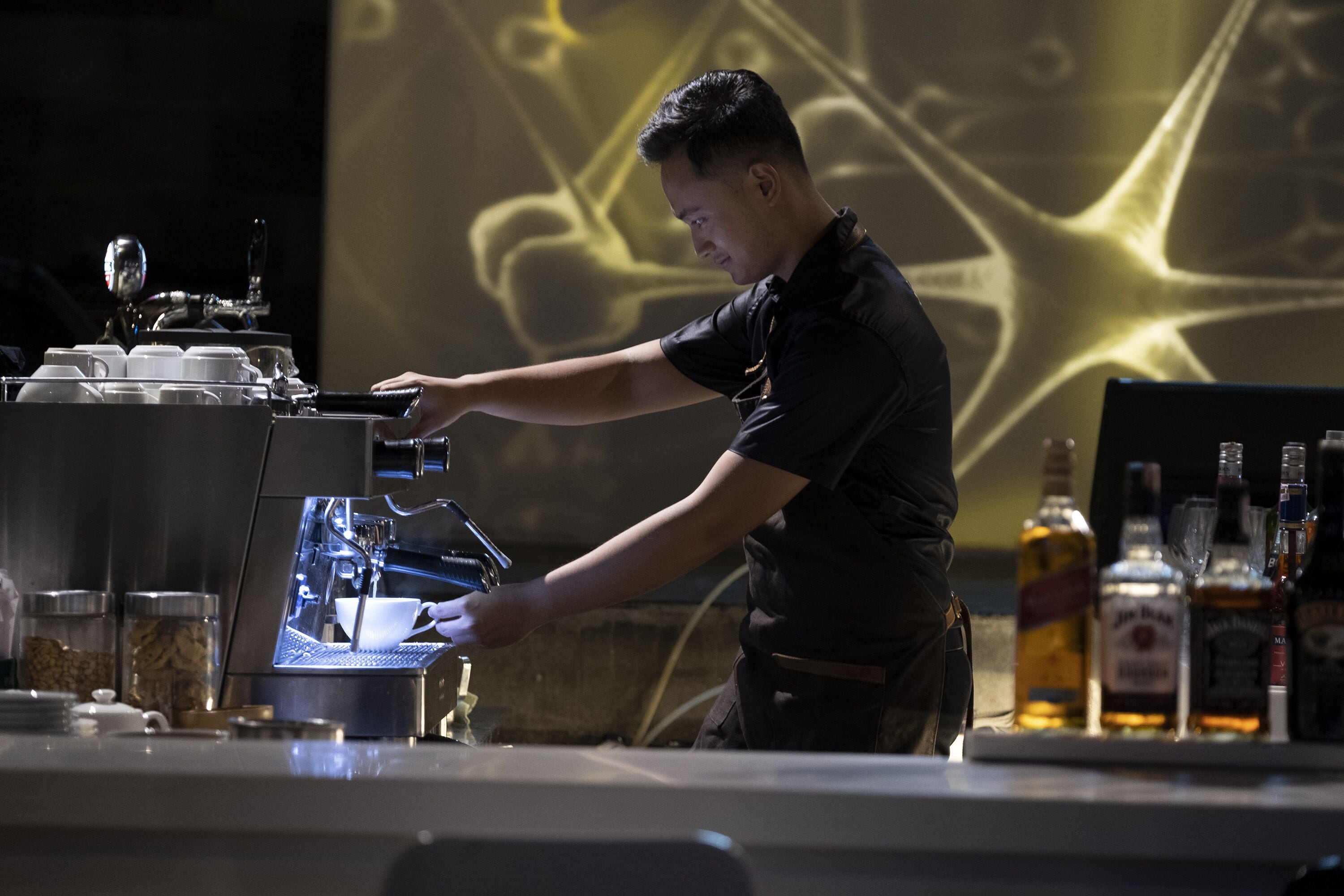 Un barista prépare une boisson à l’aide d’une machine à café, avec une projection visible en arrière-plan.