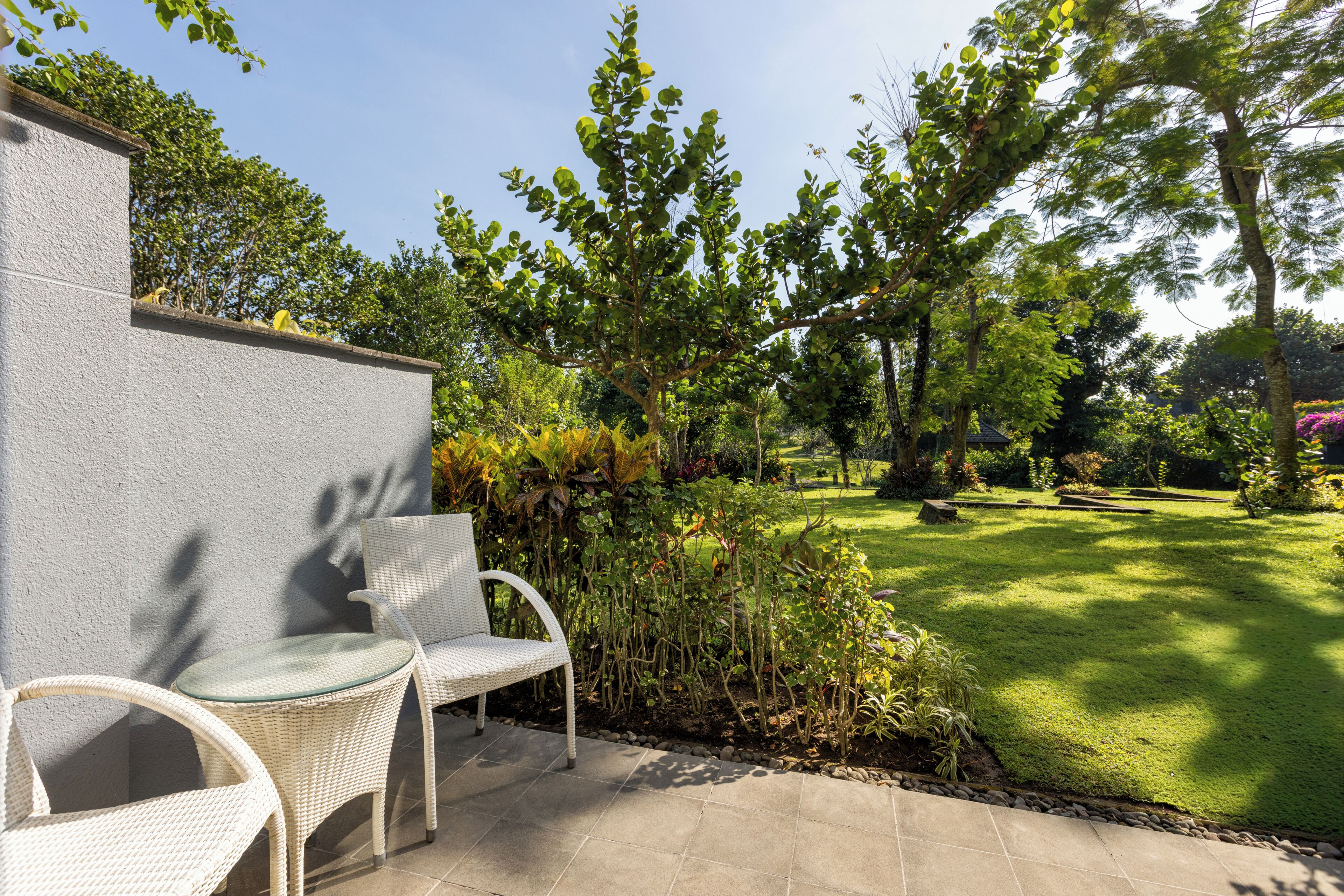 A patio with two wicker chairs, a stone wall, and a well-maintained lawn with trees and shrubs.