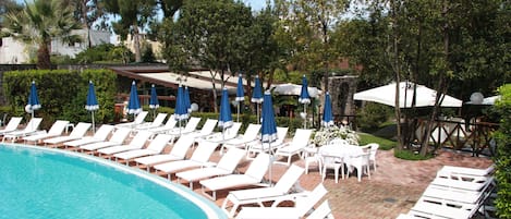 A poolside area with white lounge chairs and blue umbrellas, surrounded by trees and a brick patio.
