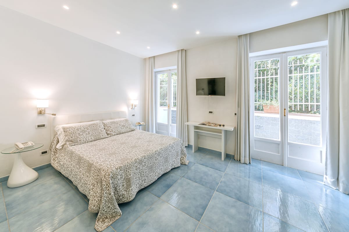 A modern bedroom with a large bed, a desk, a TV, and a view of the outdoors through large windows.