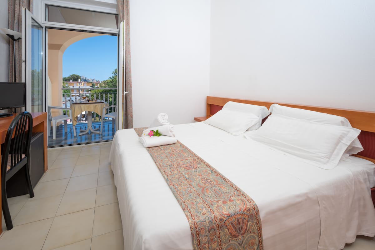 A hotel room with a bed, a desk, a chair, a table, and a balcony with a view of buildings and trees.