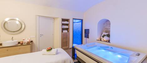 A hotel room with a large bathtub, a round mirror, a vanity with toiletries, and a door leading to a shower area.