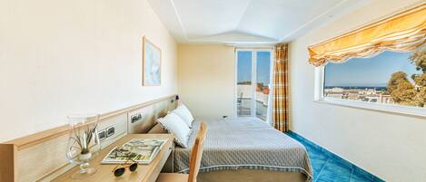 A compact bedroom with a bed, a desk with a chair, a window with curtains, and a view of the city.