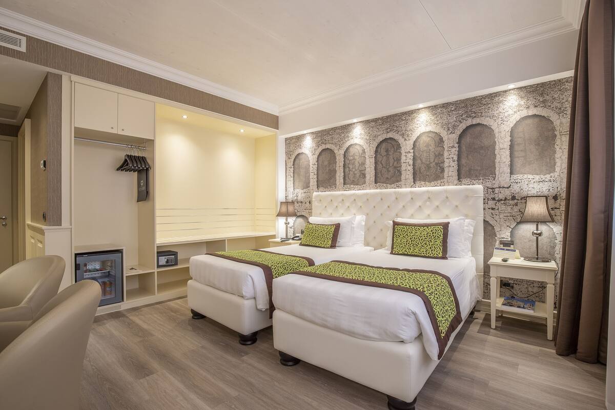 Habitación de hotel con dos camas, un armario, un espacio de descanso y una mesita de noche con lámpara.