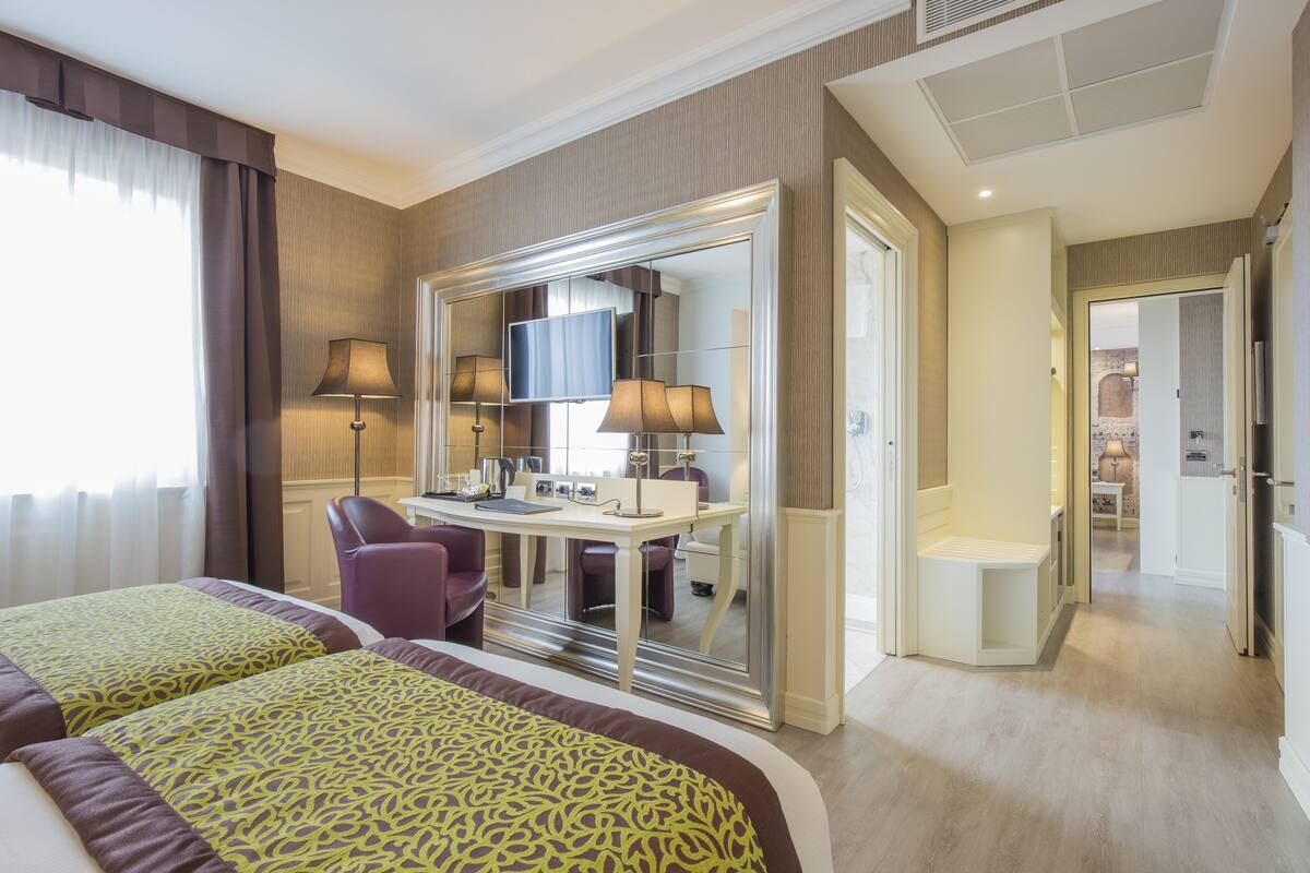 Habitación de hotel con dos camas, un escritorio, una silla, un televisor y un baño visible a través de una puerta de vidrio.