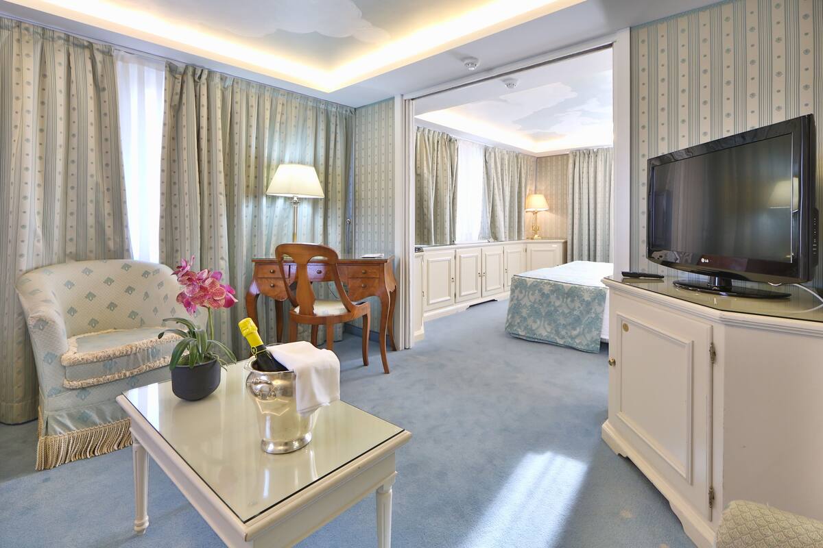 A hotel room with a sofa, a table with a bottle and flowers, a TV, and a bed.