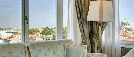 A room with a sofa and armchairs, large windows with curtains, and a lamp.