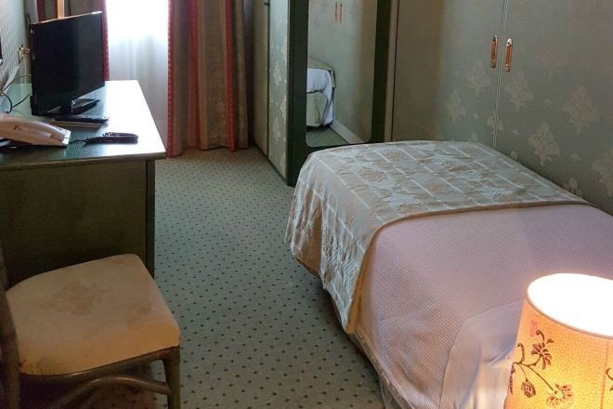 A hotel room with a bed, a desk with a computer, a chair, and a lamp.