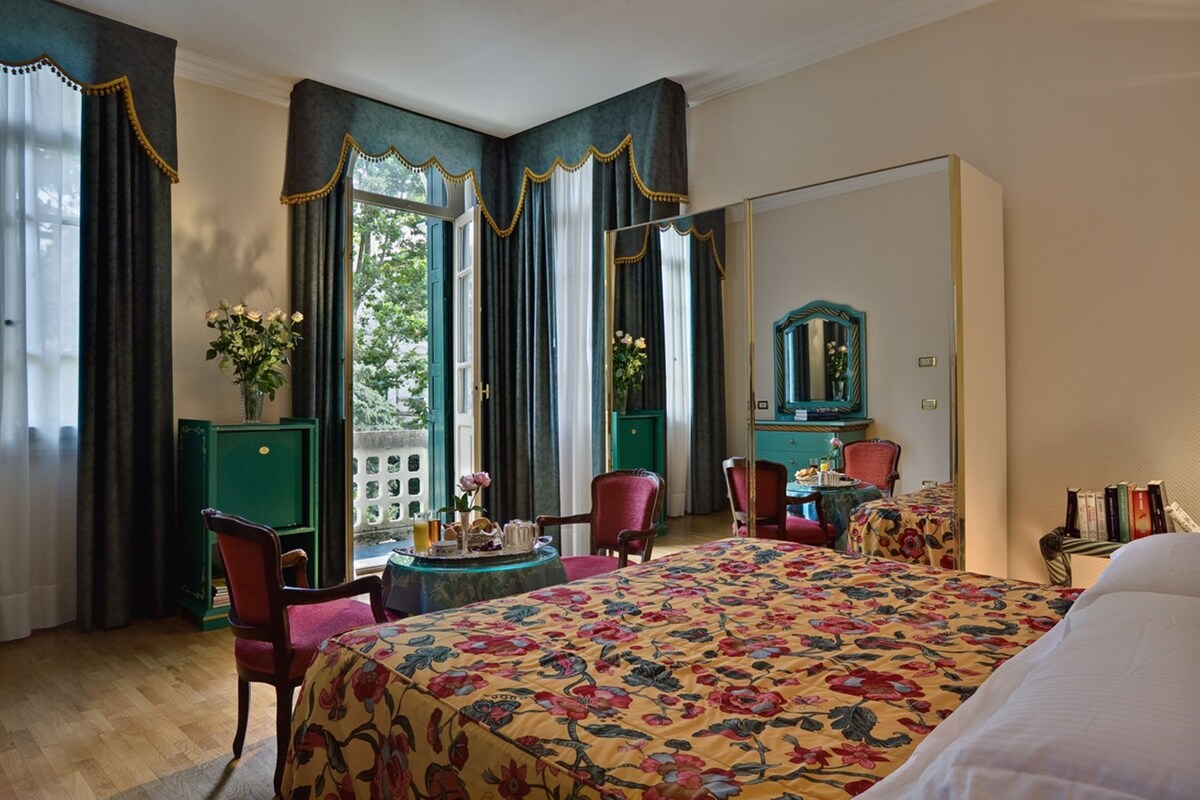 A hotel room with a large bed, two chairs, a small table with a vase of flowers, a mirror, and a green cabinet.