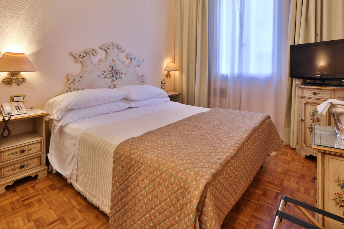 A hotel room with a bed, a television, a telephone, and a cabinet with a bottle and glasses.