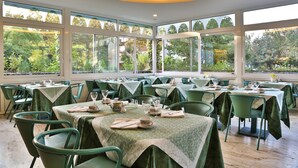 Un restaurante con varias mesas y sillas, amplias ventanas que dan a zonas verdes y un techo con una lámpara circular.
