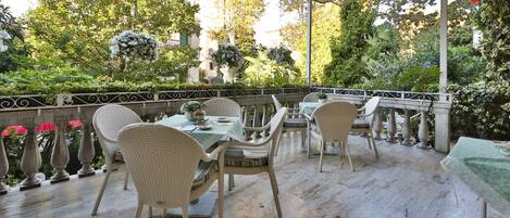 Un patio al aire libre con mobiliario de mimbre y una mesa dispuesta para comer, rodeado de vegetación exuberante y una barandilla decorativa.