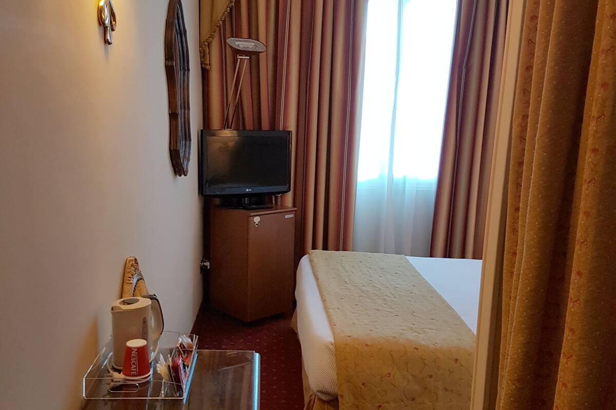 A hotel room with a bed, a desk with a coffee machine and a cup, a TV on a cabinet, and curtains.