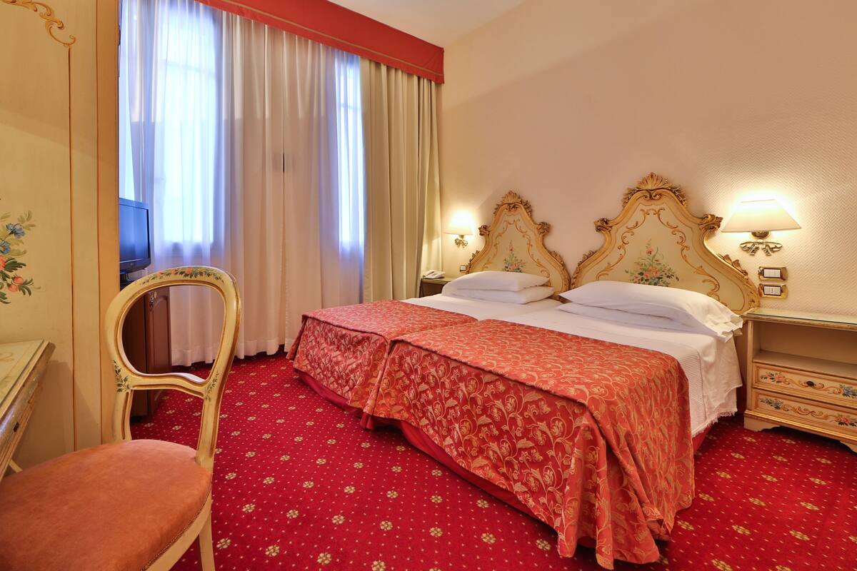 A hotel room with a double bed, a desk with a chair, a TV, and a window with curtains.