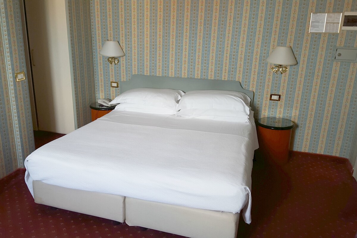 A hotel room with a bed, two bedside lamps, a nightstand, and a red carpeted floor.
