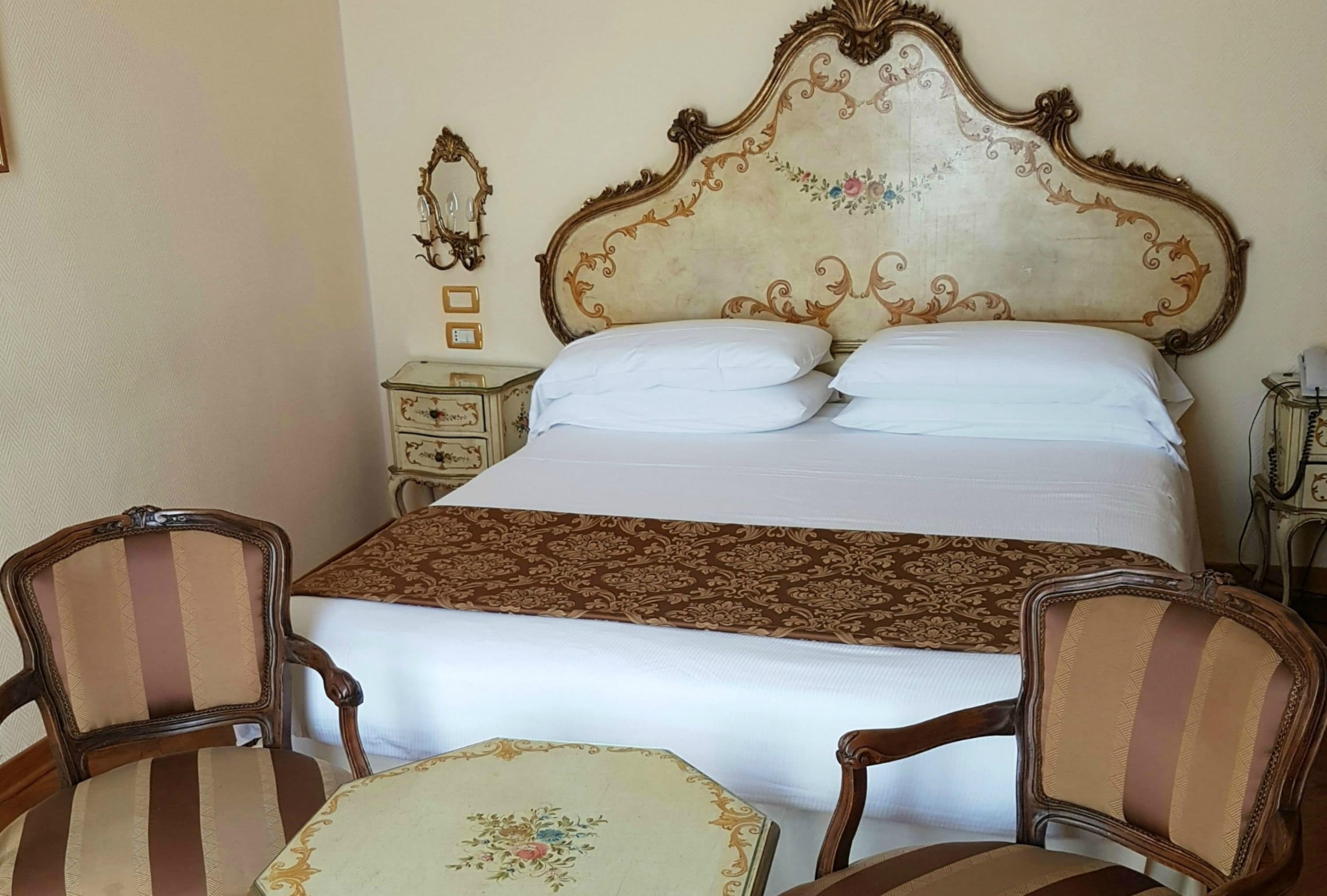 Un dormitorio con una cama grande, dos sillas, una mesita de noche y un espejo montado en la pared.