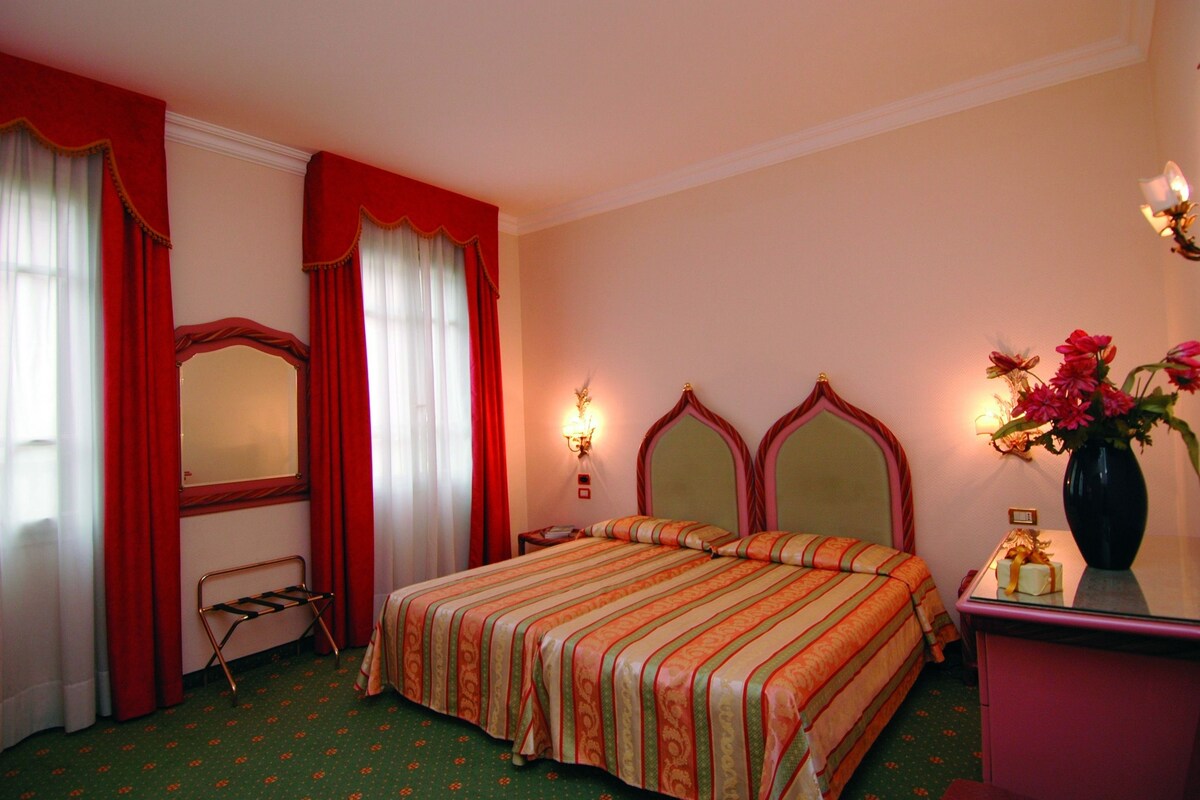 A hotel room with a bed, a vanity with a mirror, a nightstand, and a vase with flowers.