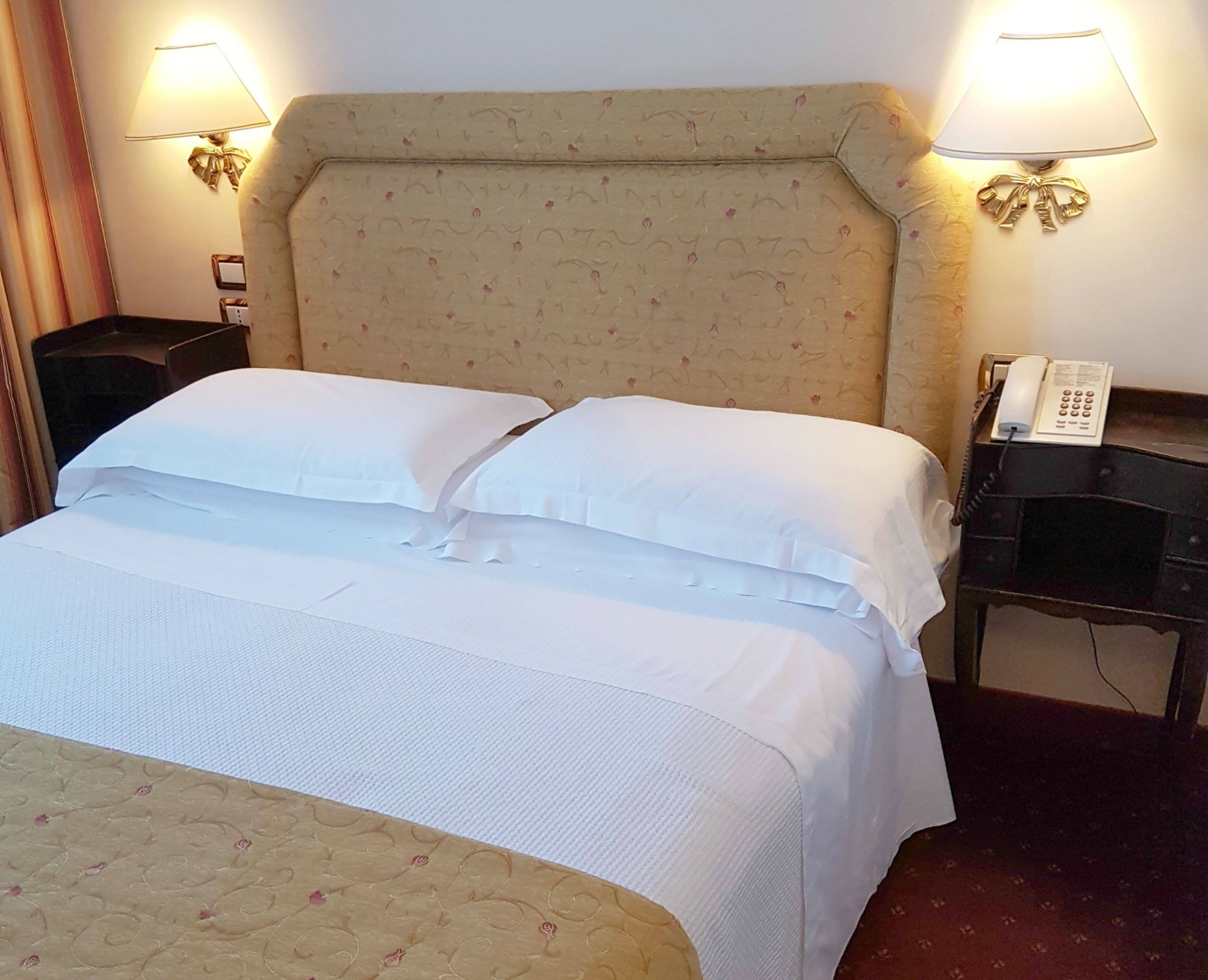 A neatly made bed with white linens, a headboard with a floral pattern, two bedside lamps, and a telephone on a nightstand.