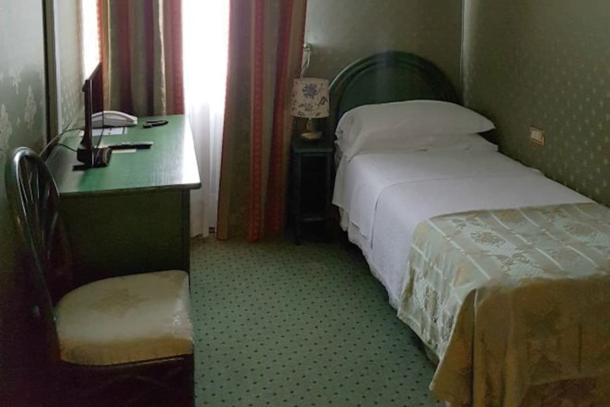 Una habitación pequeña con cama individual, escritorio y silla, mesita de noche con lámpara, y ventana con cortinas.