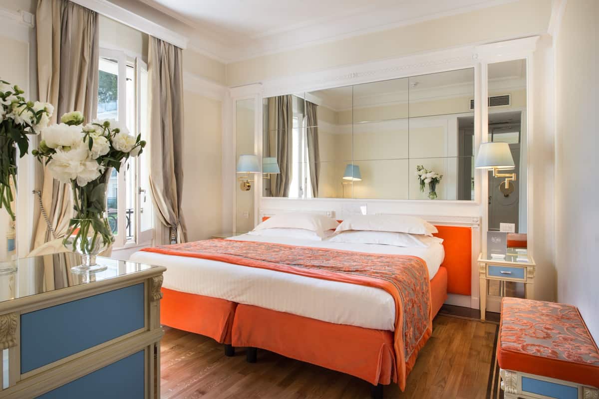 Habitación de hotel con una cama grande, una mesita de noche con lámpara, un espejo y una ventana con cortinas.