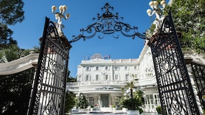 Una entrada imponente con puertas de hierro ornamentadas, una fuente y un edificio con balcones y banderas.