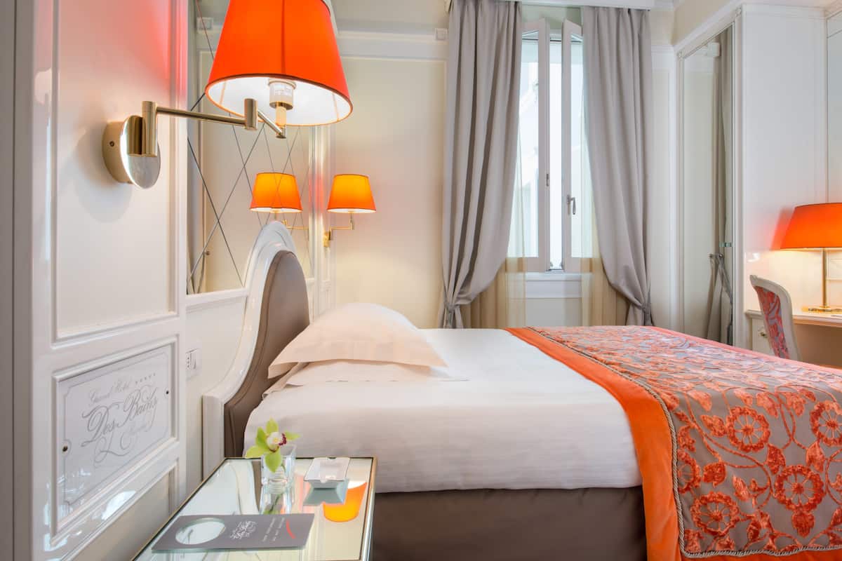 Una habitación de hotel con cama, una mesita de vidrio con lámpara y una ventana con cortinas.