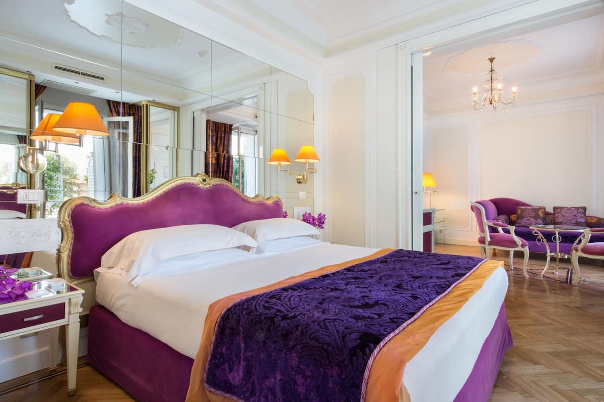Una cama grande con cabecera morada, una mesita de noche y un espejo. En el fondo, hay una silla y una mesa con una lámpara.