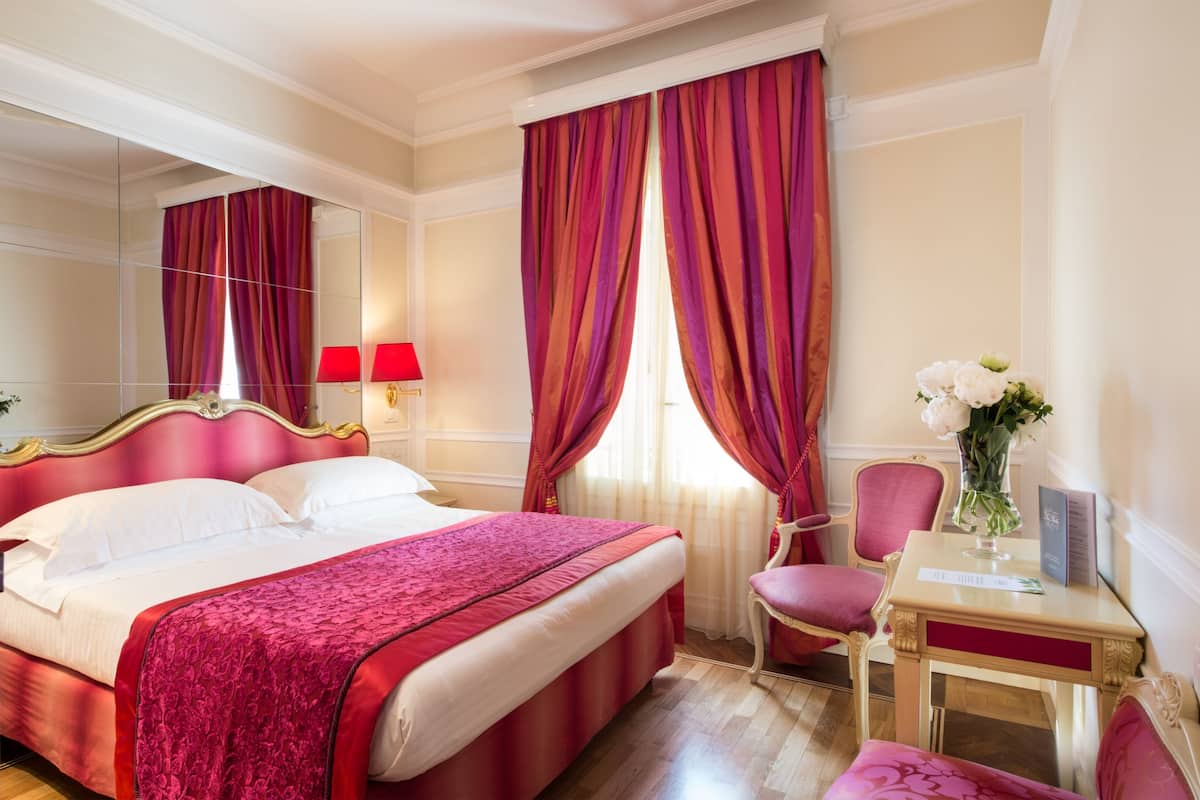 Habitación de hotel con una cama grande, cortinas rojas, un espejo, una silla y un florero con flores.