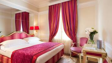 Quarto de hotel com uma cama grande, cortinas vermelhas, um espelho, uma cadeira e um vaso com flores.