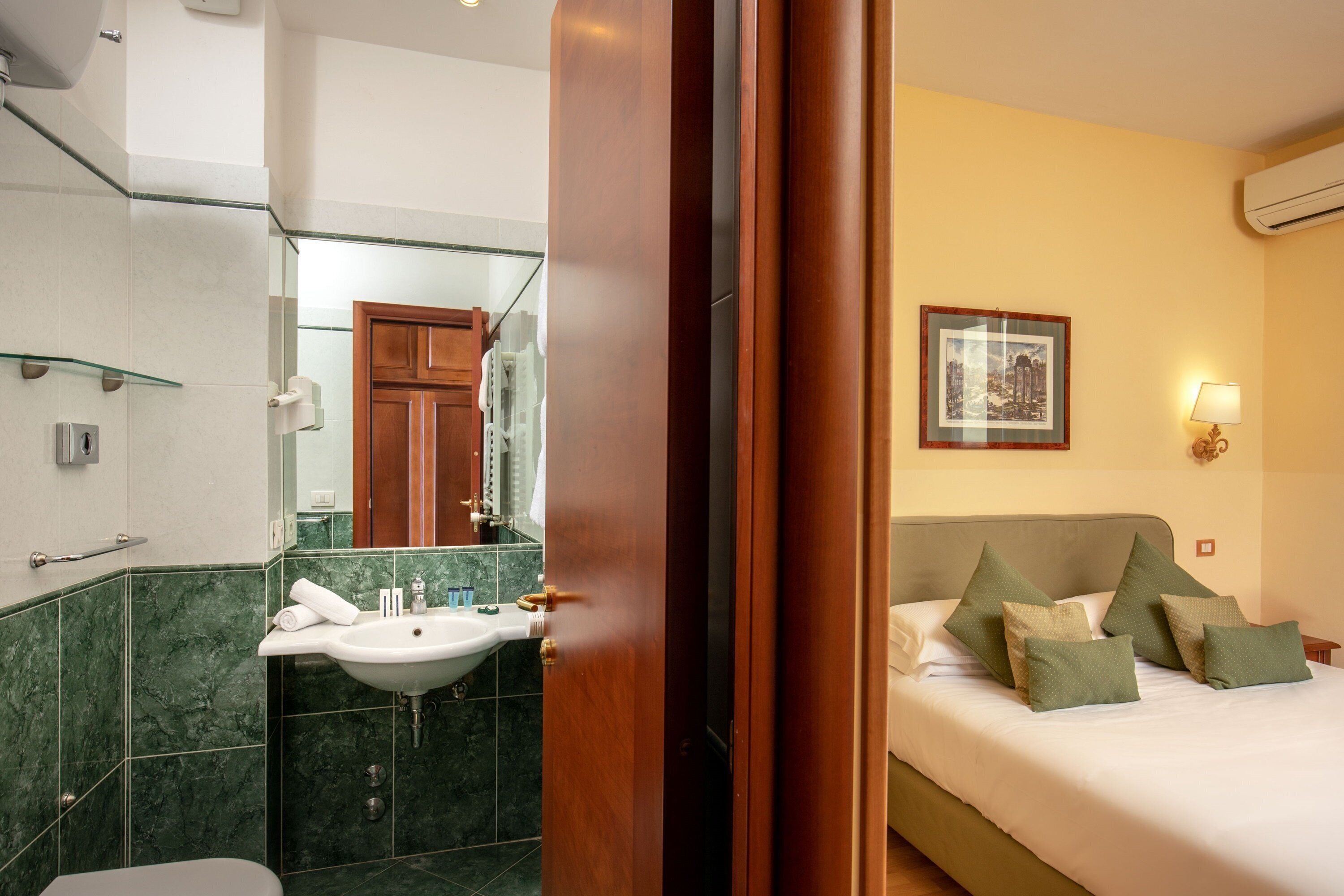 A hotel room with a bathroom featuring a sink, mirror, and green-tiled walls. The room has a bed with green pillows and a framed picture on the wall.