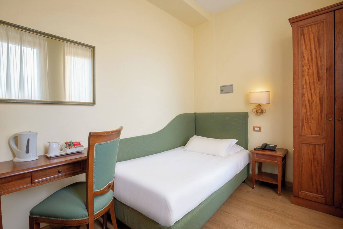 Hotellihuone, jossa on sänky, työpöytä tuolilla, vaatekaappi ja yöpöytä lampulla.
