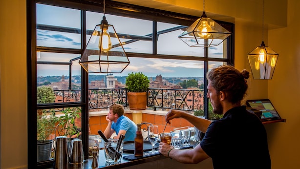 Zona de bar con vistas a un paisaje urbano a través de amplios ventanales, una planta en maceta y lámparas colgantes.