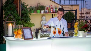 Un bartender preparando una bebida en un bar al aire libre con una mesa blanca, rodeado de plantas en macetas, una vela y varias botellas.
