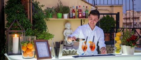 A bartender preparing a drink at an outdoor bar with a white table, surrounded by potted plants, a candle, and various bottles.