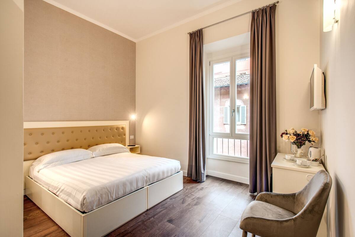 A hotel room with a bed, a chair, a small table with a vase of flowers, and a window with curtains.