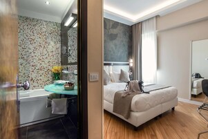 A modern hotel room with a glass-enclosed bathroom, a bed with a beige duvet, and a wooden floor.