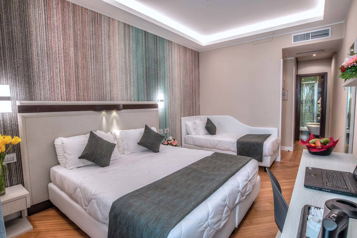 A modern hotel room with a large bed, a smaller sofa, a desk with a laptop, and a decorative vase with flowers.