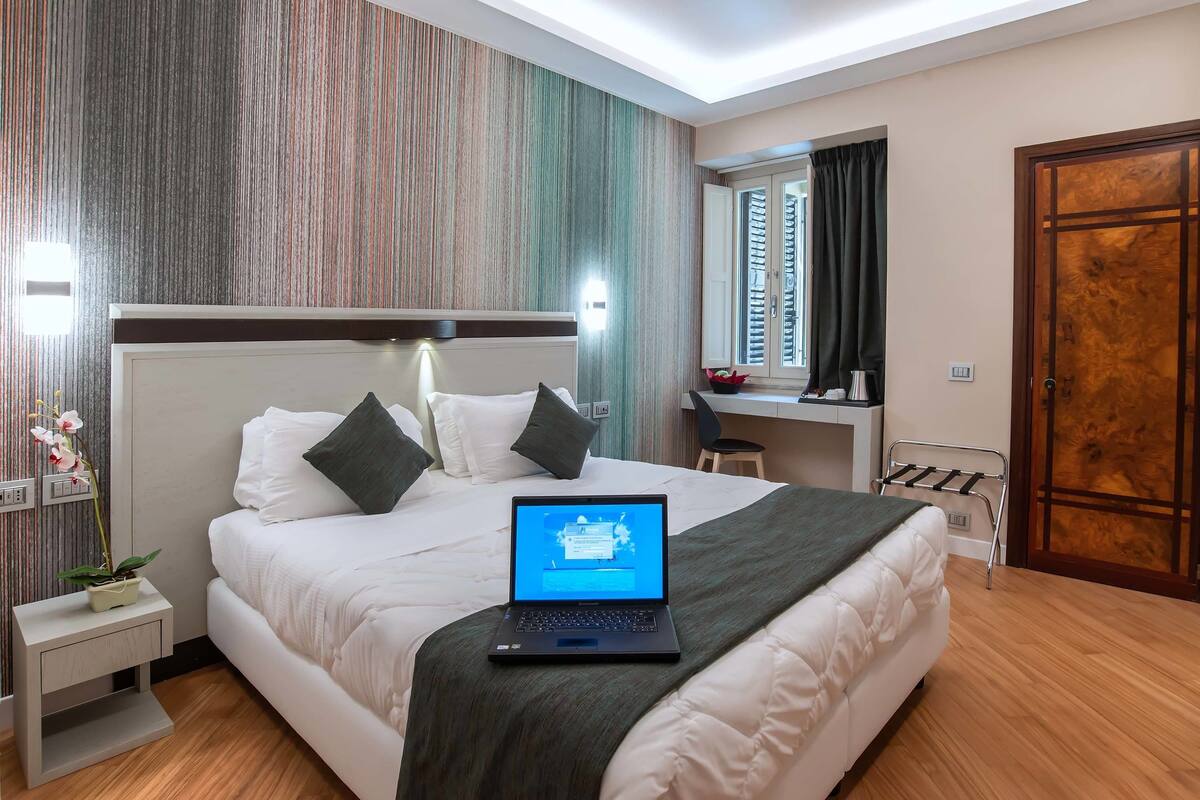 A modern hotel room with a neatly made bed, a laptop on the bed, a small bedside table with a plant, a desk with a chair, and a large wooden door.