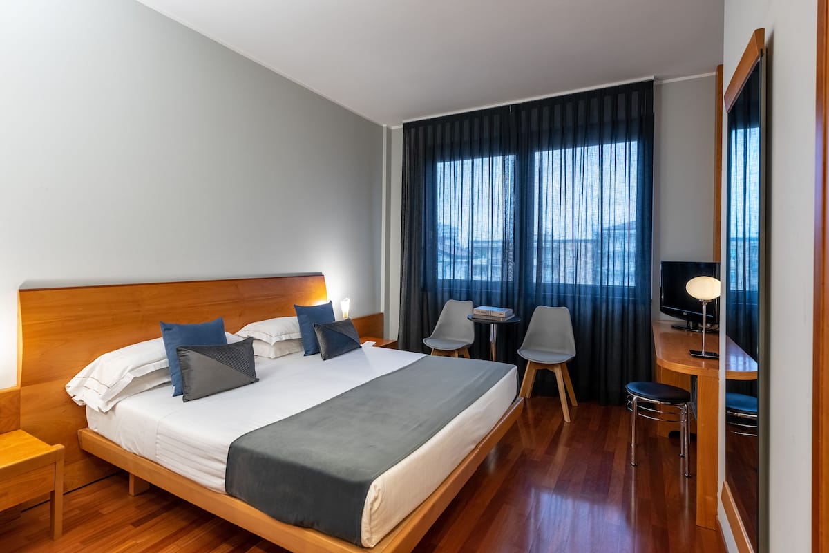 A hotel room with a large bed, bedside tables, a desk with a chair, a TV, and a view of the city through the window.