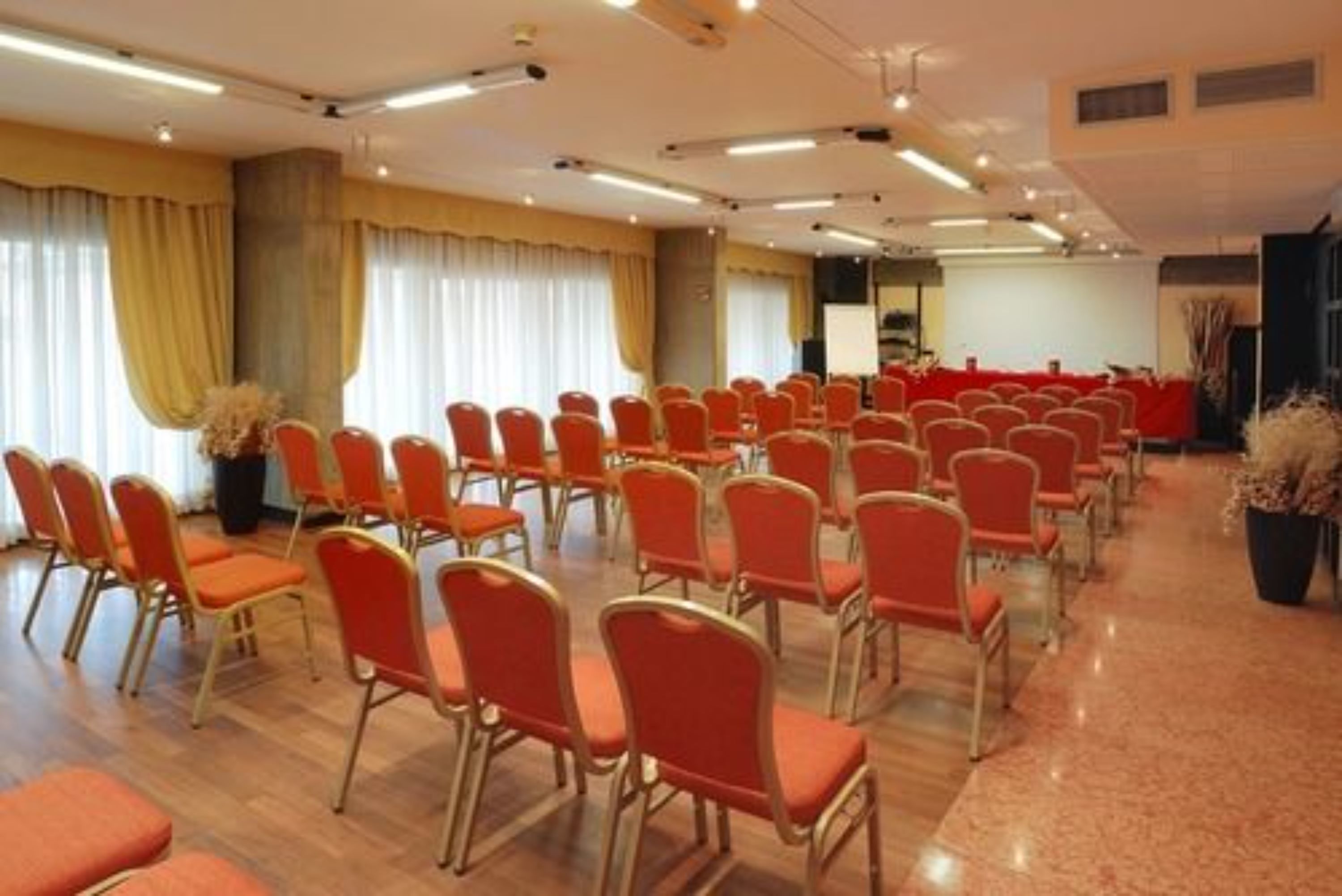 A conference room with rows of orange chairs, a stage with a whiteboard, and large windows with curtains.