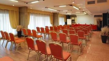 Un salón de conferencias con filas de sillas naranjas, un escenario con una pizarra y ventanas grandes con cortinas.