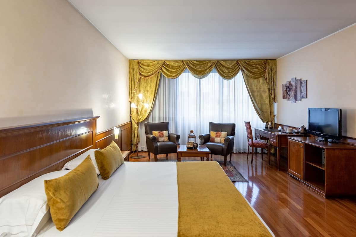 A hotel room with a large bed, two armchairs, a desk, a television, and a window with curtains.