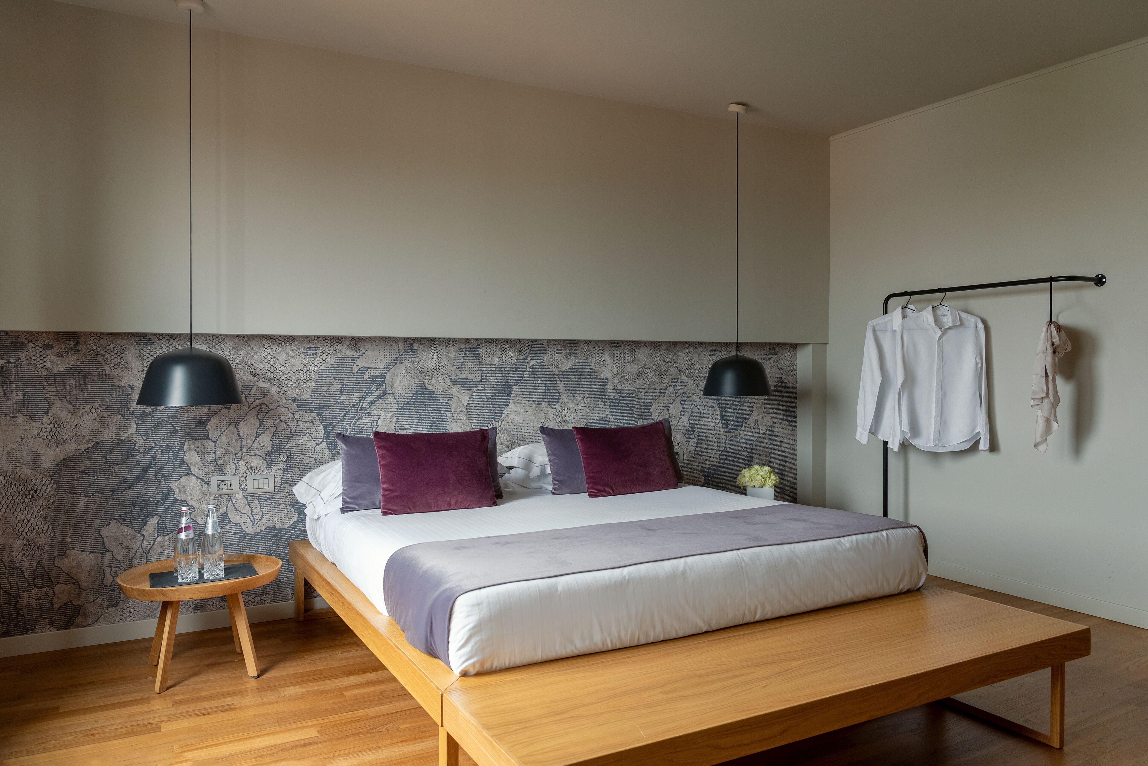 A modern bedroom with a wooden bed, two black pendant lamps, a small round table with bottles, and a patterned wallpaper.